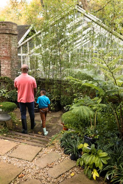 Senior African American man and his grandson walking through a lush garden nursery. Ideal for concepts related to family bonding, intergenerational relationships, gardening, nature, and outdoor activities. Useful for articles, advertisements, and educational materials focusing on family, horticulture, and leisure activities.