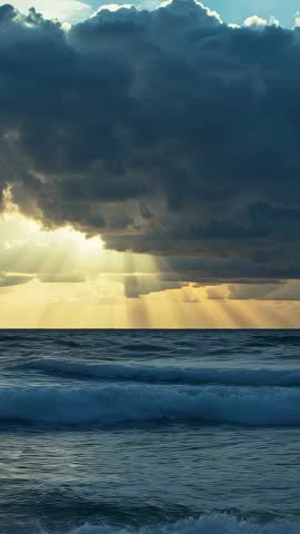 Vertical ocean sunset with sunbeams piercing storm clouds over rolling swells and shore foam
