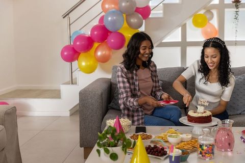 Friends cutting 18th birthday cake on couch with balloons and party snacks