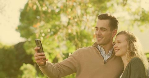 Smiling couple taking selfie in sunlit park during golden hour enjoying outdoor moment