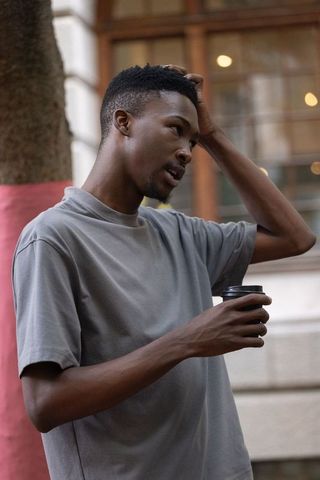 Man leaning on city sidewalk holding coffee cup touching hair, candid urban lifestyle