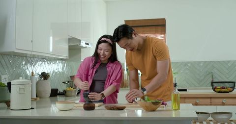 Young asian couple preparing fresh salad and grinding spices together in modern kitchen