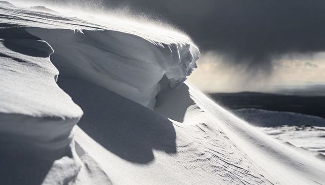 Wind-carved snow cornice curving over alpine ridge with blowing powder and storm clouds