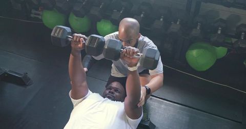 African man pressing heavy dumbbells on bench with trainer spotting and coaching