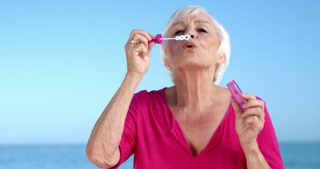 Senior woman enjoying a sunny day blowing bubbles by the ocean. This image is perfect for concepts related to age positivity, enjoying life, relaxation, and outdoor activities for senior citizens. Ideal for social media, health and wellness articles, and advertising for elder care programs.