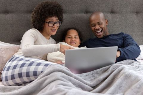 Multiracial family relaxing in bed watching laptop, laughing together under cozy blanket