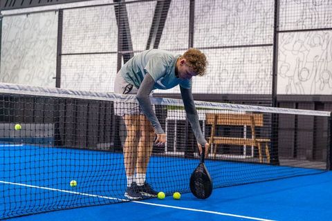 Young male athlete bending over padel net collecting balls on blue court