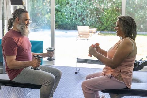 Senior couple working out with dumbbells on benches near glass doors in bright home gym