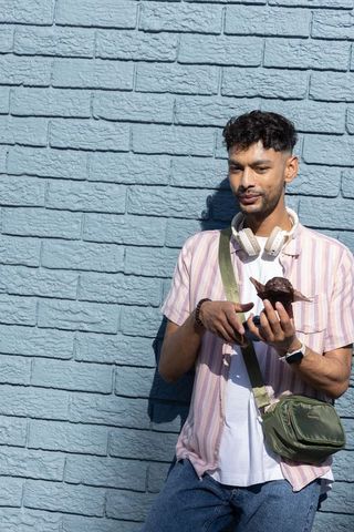 Indian man examining turtle figurine by sunlit light-blue brick wall with headphones