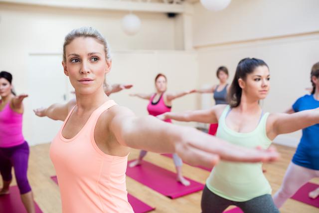 Group of women participating in a stretching exercise at a well-lit fitness studio. They are wearing sportswear and demonstrating a synchronized activity, likely part of a yoga or fitness class. Perfect for promoting fitness studios, wellness programs, group exercise classes, or healthy lifestyle campaigns.