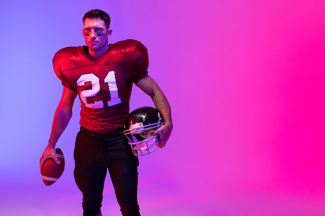 Caucasian male american football player holding helmet and ball, in pink and blue light, copy space. Athlete, sport, competition, confidence and fitness concept.