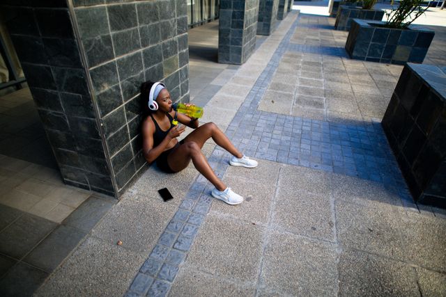 African American woman wearing sportswear and headphones, sitting on pavement against a wall, drinking water after an outdoor workout. Ideal for use in fitness blogs, health and wellness articles, sportswear advertisements, and lifestyle magazines promoting active and healthy living.