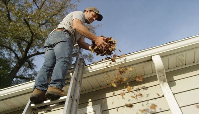 Homeowner cleaning gutters from ladder removing autumn leaves for roofline maintenance