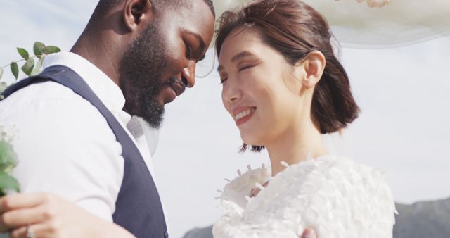 Interracial couple sharing a tender moment on their wedding day as they embrace outdoors, with smiles on their faces and a soft sky backdrop. This image is perfect for wedding invitations, marriage blogs, love and relationship websites, and promotional materials for wedding services.