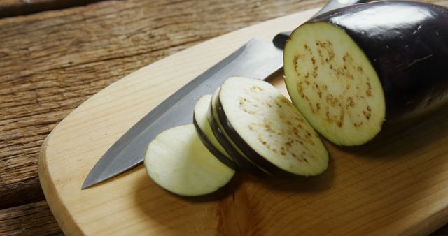 Sliced eggplant arranged on a wooden cutting board next to a knife. Ideal for food preparation, recipes, vegetarian and vegan meal planning, kitchen scenes, culinary blogs, and advertisements promoting fresh vegetables.