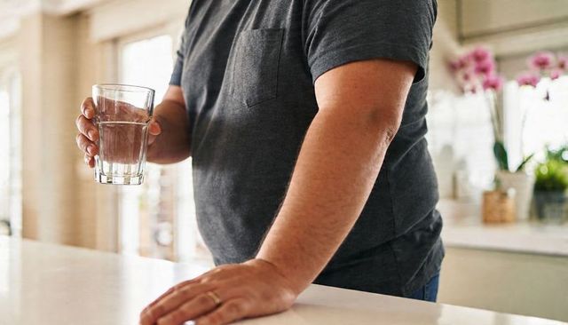 Man holding glass of water at sunlit kitchen counter showing wedding ring close-up for home hydratio