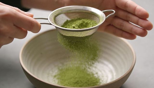 Hands sifting vibrant matcha powder through fine mesh sieve into ceramic bowl