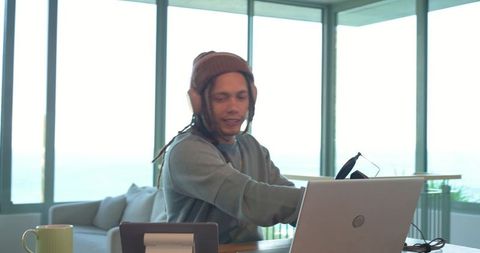 Mid-adult man working remotely in oceanview home office wearing knit beanie and headphones