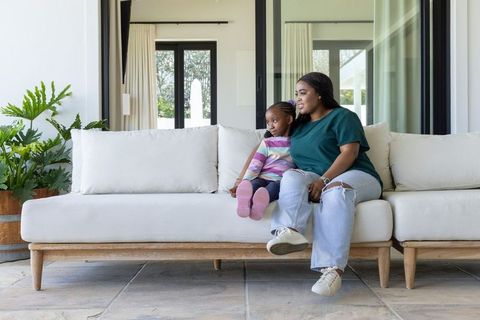 African american mother and daughter relaxing on white sofa by glass doors in sunlit home