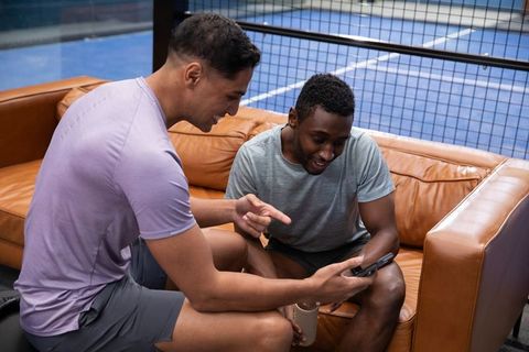 Two men sharing smartphone on leather sofa beside indoor tennis court