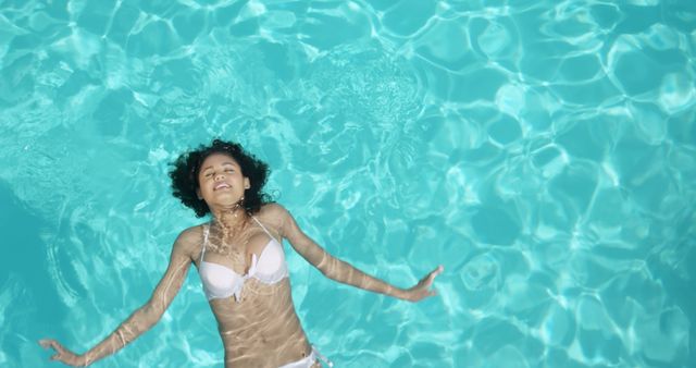 Young woman is relaxing and floating in a swimming pool wearing a white bikini. She appears to be enjoying the water and the warm weather. This image can be used for summer vacation promotions, travel advertisements, lifestyle blogs, and wellness articles.