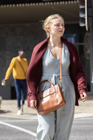 Young woman walking city street wearing blue jumpsuit, burgundy cardigan carrying tan handbag