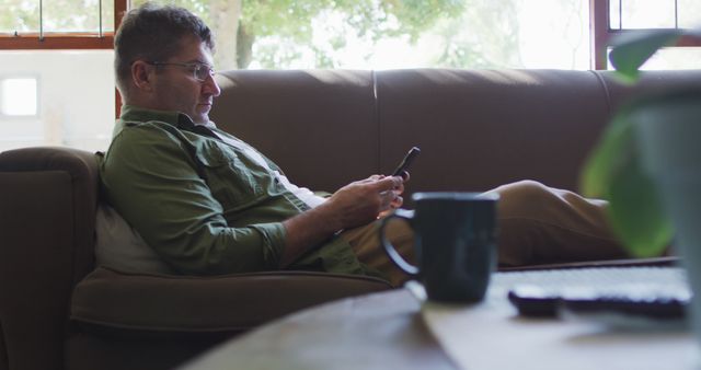 This depicts a man relaxing on a sofa, engrossed in using his smartphone. The relaxed posture and daylight hint at a laid-back, casual setting, perfect for use in lifestyle blogs, home decor, or technology advertisement that emphasizes comfort and modern living.