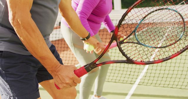 Image of midsection of biracial senior couple holding rackets on tennis court. active retirement lifestyle, senior relationship and tennis training concept.