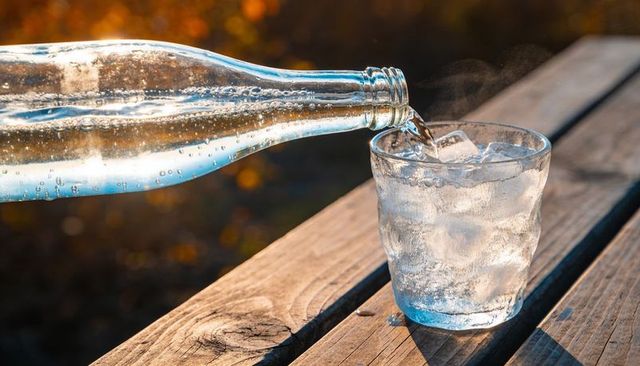 Pouring sparkling water from glass bottle into ice-filled tumbler on sunlit wooden table