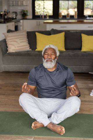 Senior african american man meditating at home on yoga mat with wrist tracker and cozy sofa