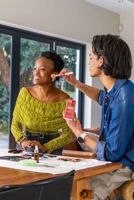 Makeup artist applying blush to smiling client holding pink palette in bright living room