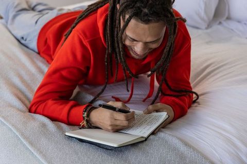 Young african american man journaling on bed wearing red hoodie and dreadlocks casual cozy