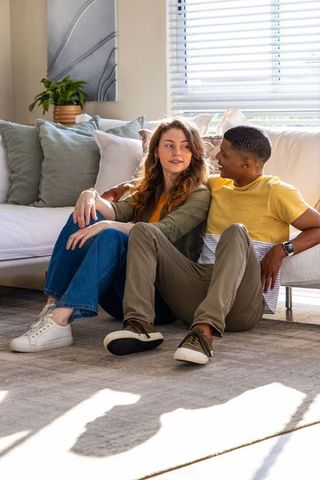 Young interracial couple sitting on rug leaning against sofa enjoying relaxed conversation