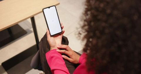 Businesswoman holding smartphone with blank notch screen sitting in pink coat near wooden table