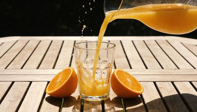 Pouring orange juice into glass with fresh orange halves on sunlit wooden deck