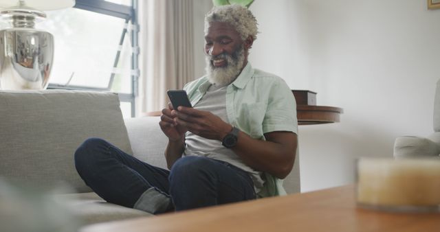 Senior man with grey hair and beard relaxing on sofa while using smartphone in modern living room. He is casually dressed in a light shirt and jeans, looking content and connected. This image is ideal for concepts like retirement, staying connected, everyday technology use, senior well-being, and comfortable home environments.