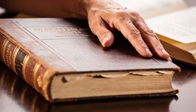 Elderly hand resting on antique leather-bound book with gilt spine, vintage study mood