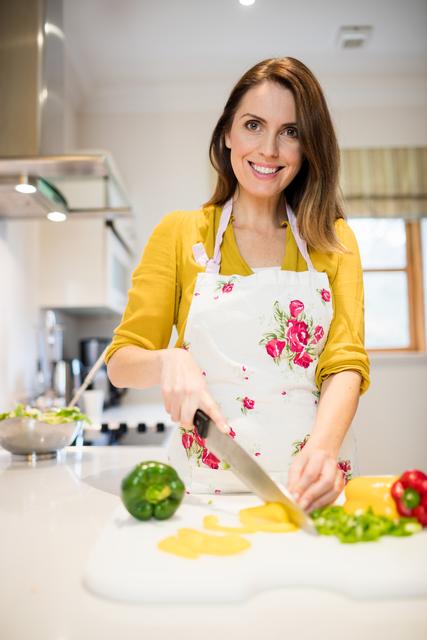 Smiling woman in modern kitchen chopping vegetables on a white cutting board. She is wearing a floral apron and a yellow shirt, creating a cheerful and inviting atmosphere. Ideal for use in articles or advertisements related to home cooking, healthy eating, lifestyle blogs, culinary tutorials, and kitchen appliance promotions.