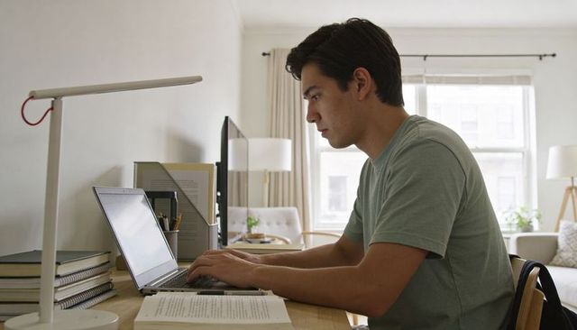 Young man working from home at minimalist living room desk typing on laptop and studying