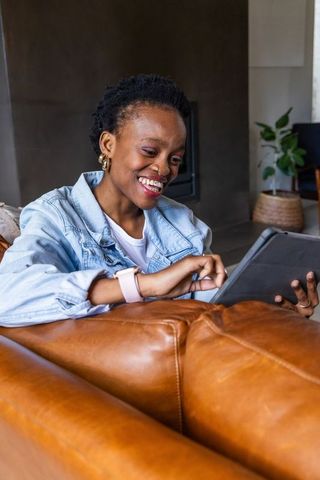 Smiling african american woman using tablet on leather couch in cozy modern living room