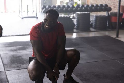 Smiling african american man crouching on gym mat wearing red shirt black shorts near weights