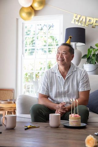 Mature asian man celebrating birthday at home wearing gold party hat with cake