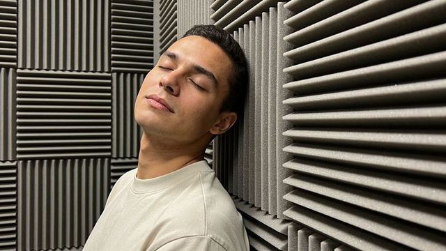 Male leaning against acoustic foam panels in recording booth, eyes closed, calm minimal scene