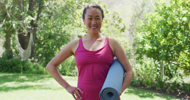 A woman is smiling and holding a rolled-up yoga mat in an outdoor park with lush greenery. This image is perfect for promoting outdoor fitness activities, yoga classes, wellness retreats, or healthy lifestyles. It can also be used for fitness blogs, health magazines, or wellness apps.