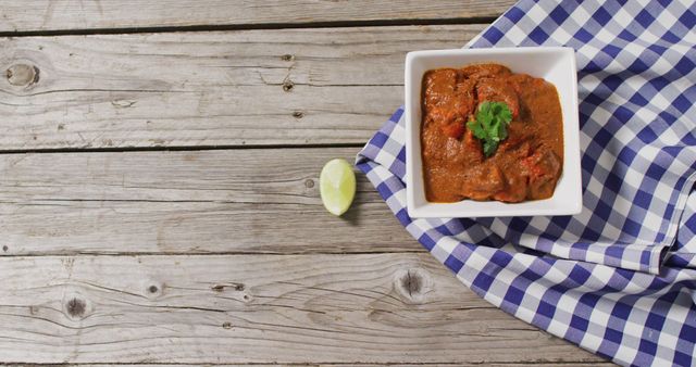 Indian chicken curry served in a white bowl garnished with cilantro on a rustic wooden table. A slice of lime is also presented beside the bowl, along with a blue checkered napkin. Ideal for use in culinary blogs, food advertising, menus, or recipe websites.