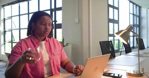 African woman working at laptop in modern coworking space holding glass and typing