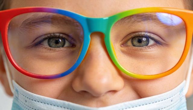Close-up child wearing rainbow eyeglasses and surgical mask showing eye reflections