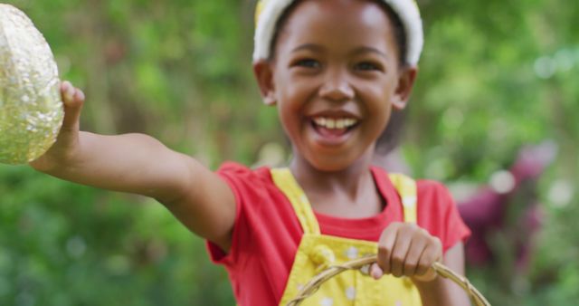 Young African American girl wearing red shirt and yellow apron, smiling and holding a woven basket outdoors. Ideal for themes related to childhood, outdoor activities, nature, happiness, and summer fun.