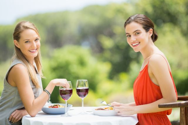 Two women are enjoying a meal together at an outdoor restaurant. They are smiling and appear to be having a pleasant conversation. The setting is bright and sunny, with greenery in the background, suggesting a summer day. This image is perfect for use in advertisements for restaurants, social gatherings, friendship themes, or lifestyle blogs.