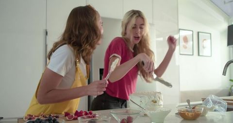 Two friends decorating cake in bright modern kitchen, laughing while frosting